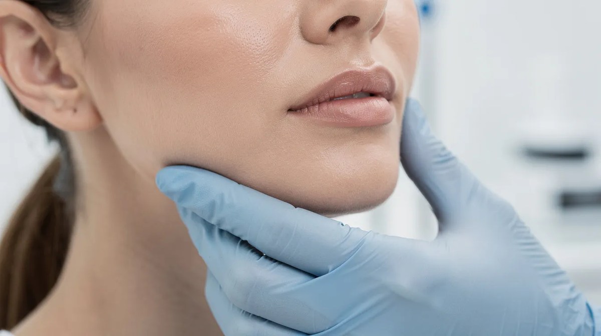 A close-up of a woman's lower face and jawline being examined by a provider wearing gloves in a clinical setting. This shows the effects of face tightening treatments, aimed at improving collagen production for a more youthful appearance.